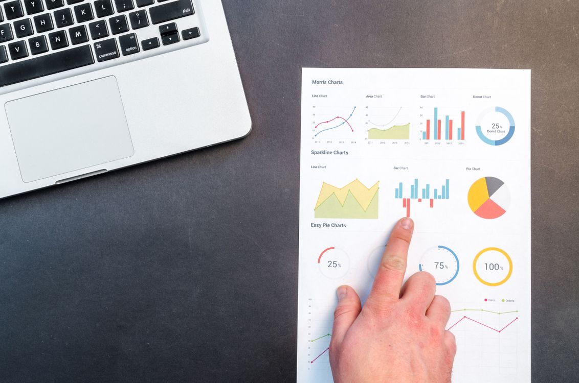 Hand pointing at colorful charts with a laptop in the background on a desk